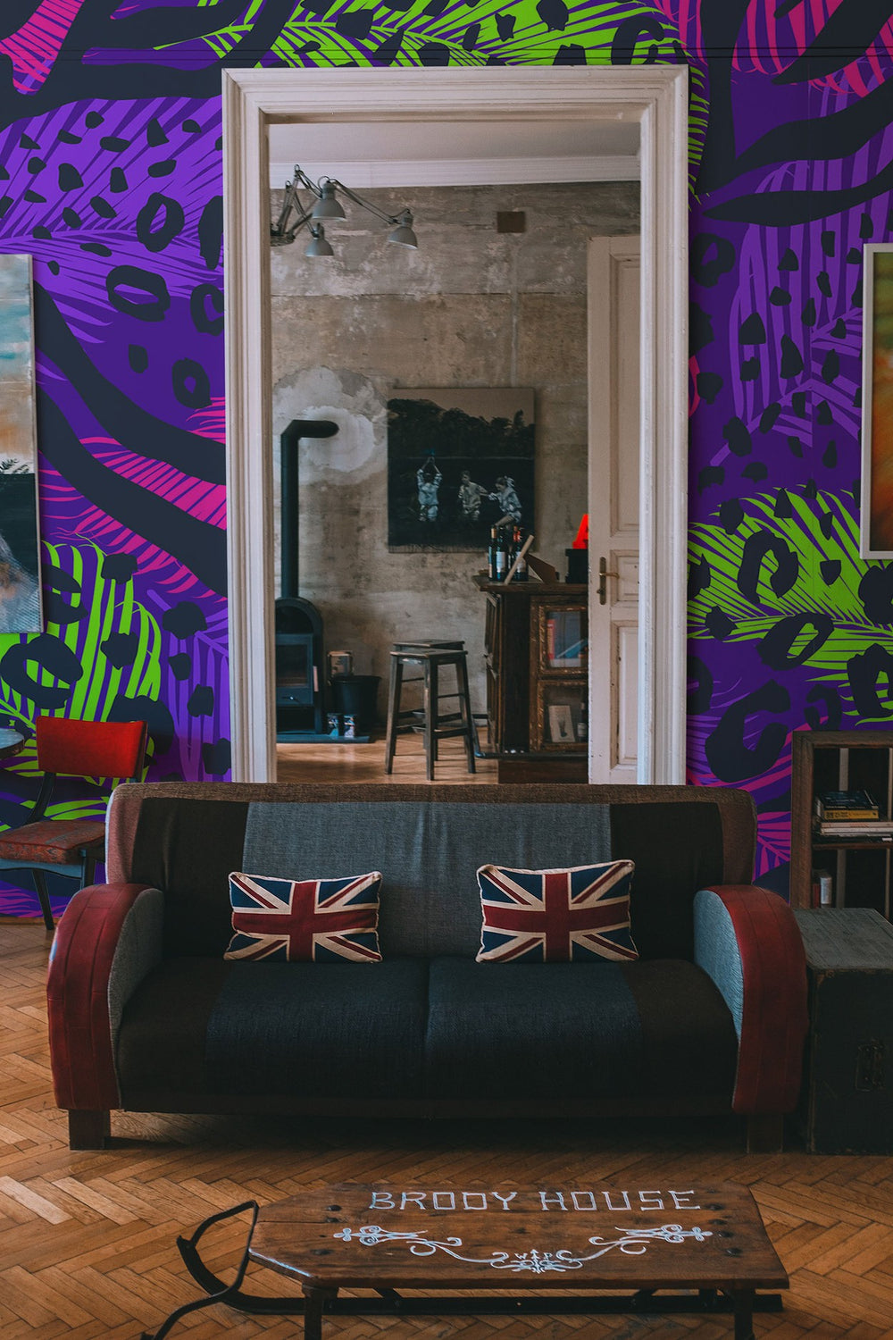 A vibrant and eclectic living room featuring a bold graphic wall mural, a dark sofa with Union Jack pillows, and a wooden coffee table with 'Brody House' text.