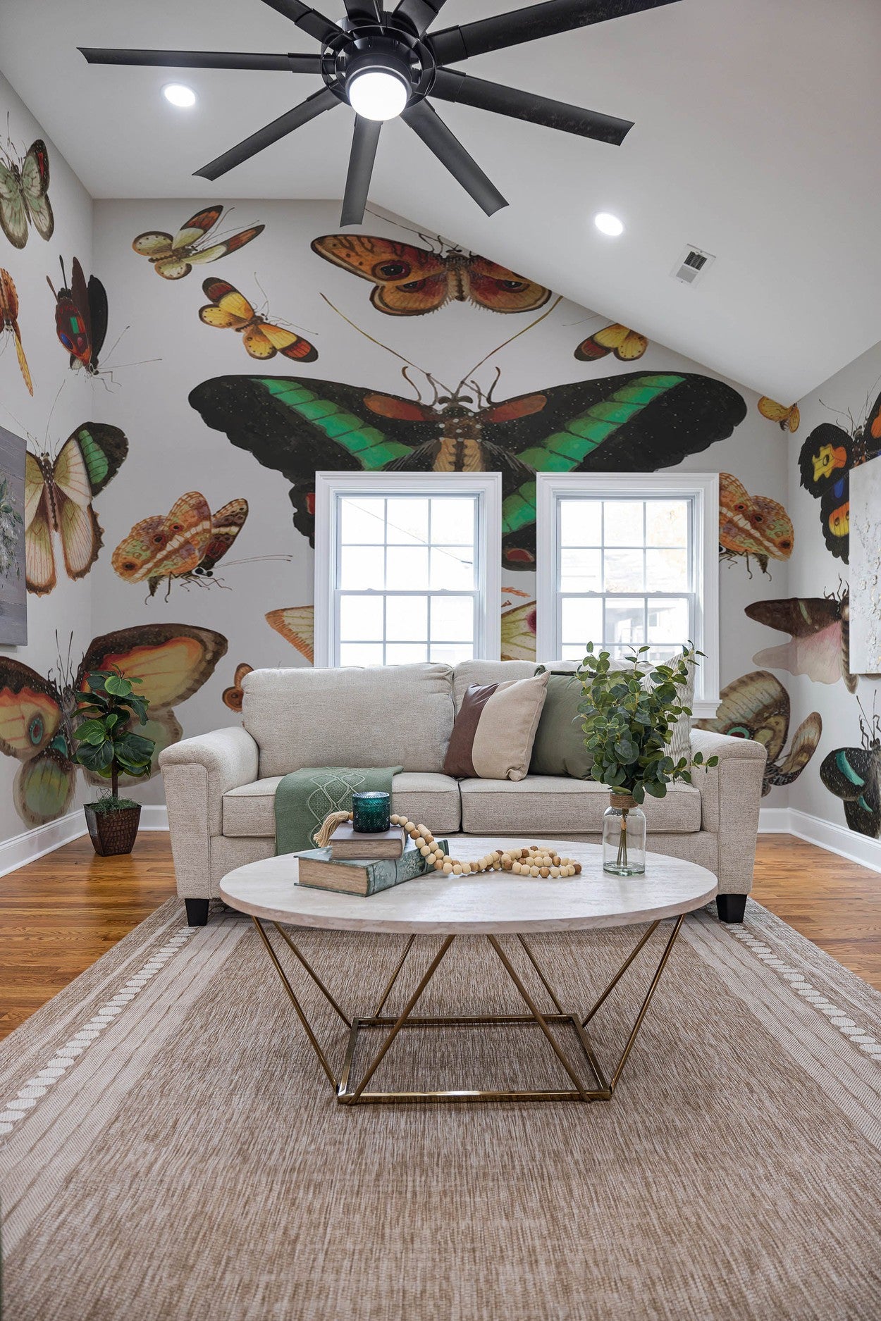 A cozy living room interior featuring a large wall mural of colorful butterflies, with a beige sofa, round coffee table, and a ceiling fan
