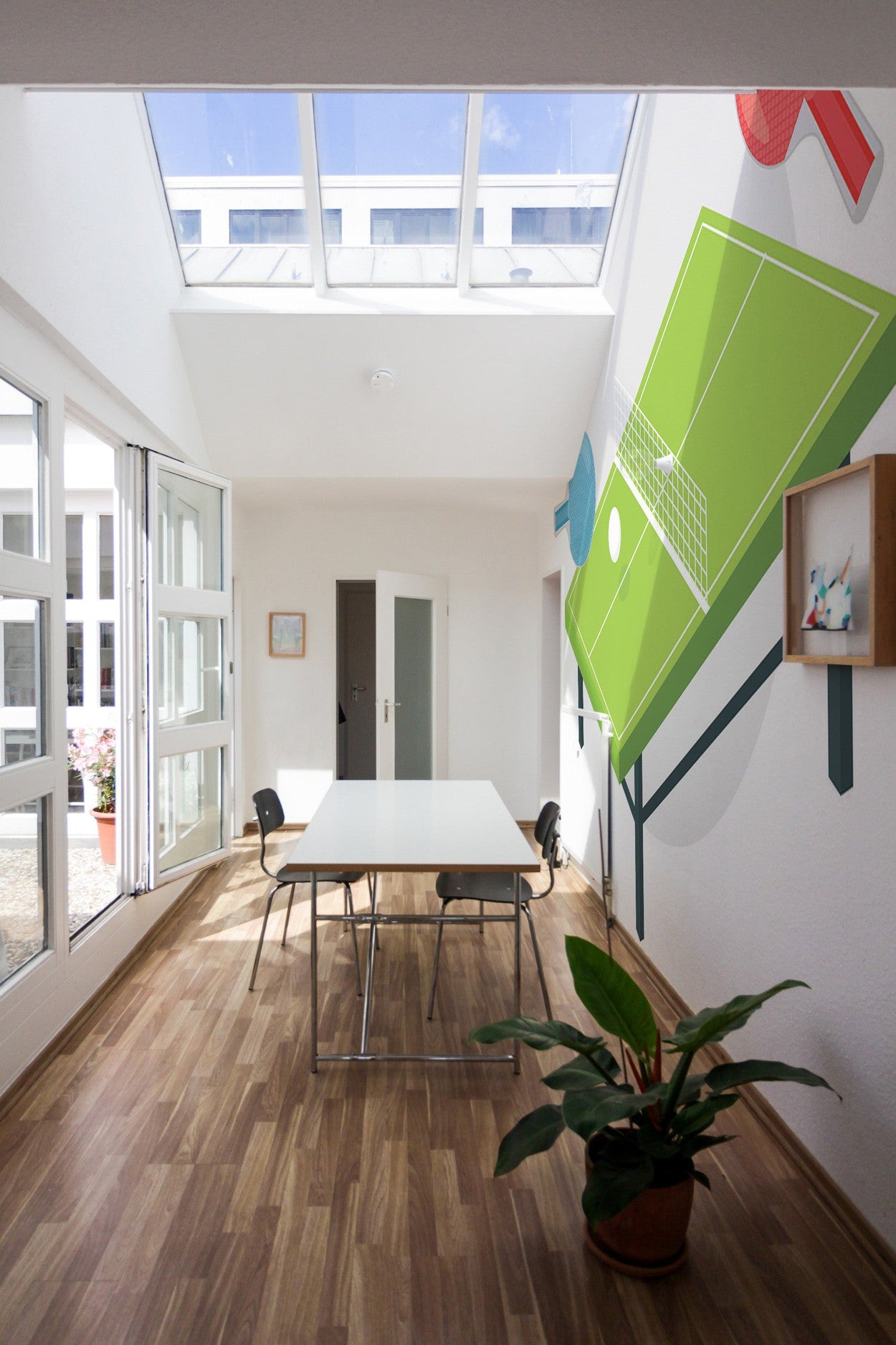 Bright room with skylight windows, wooden flooring, a simple table and chairs, and a large wall mural of a tennis court.