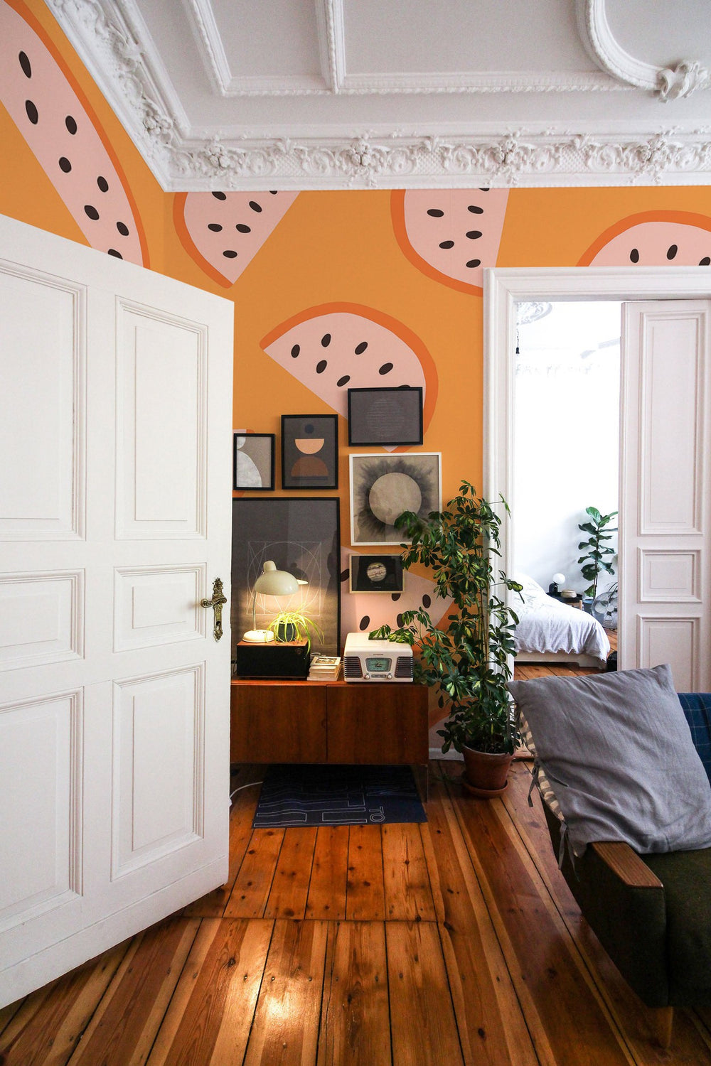 A cozy living room interior with a geometric wall mural in orange and pink tones, featuring elegant white doors, wooden flooring, and a display cabinet with decorative items.