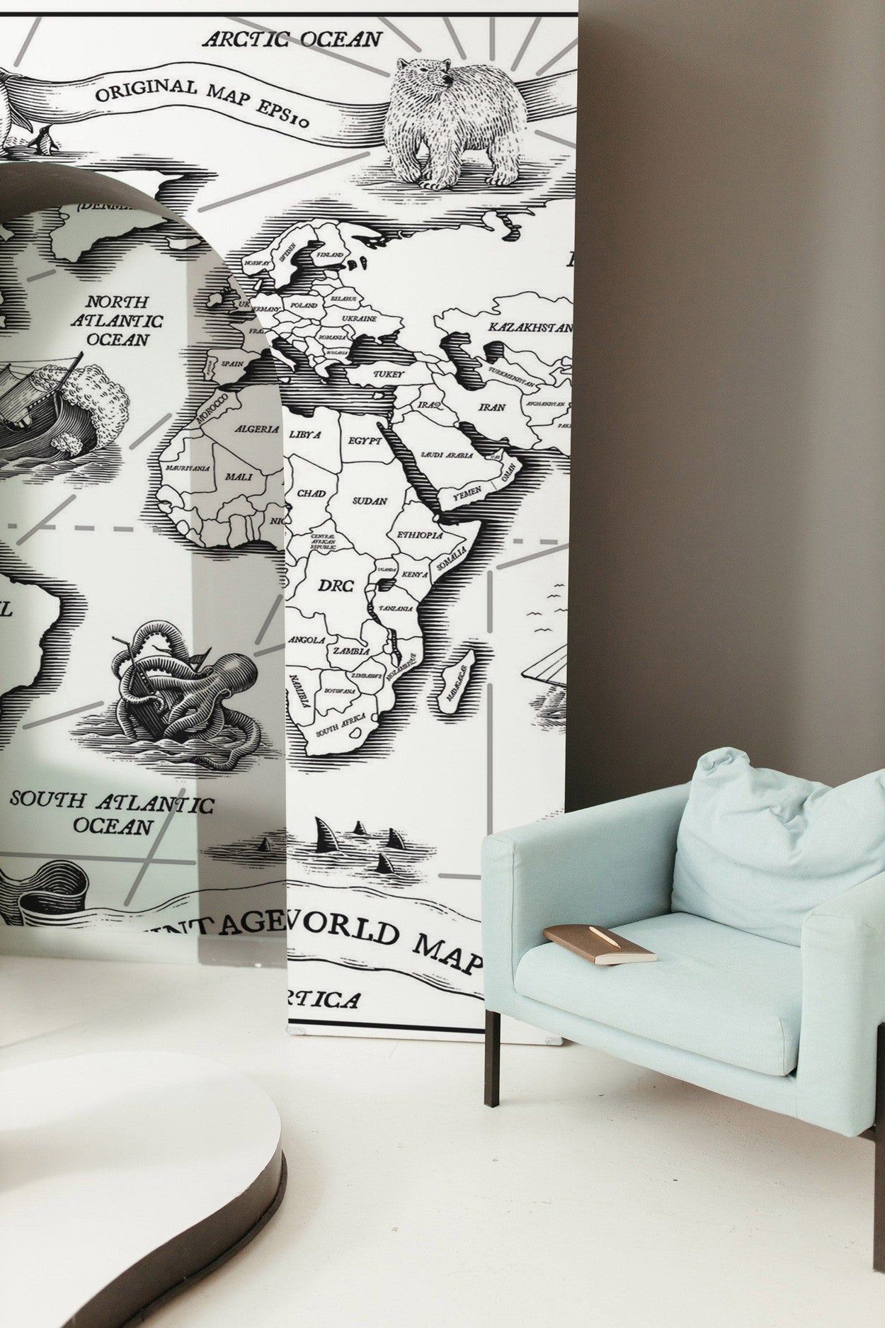 A stylish interior featuring a wall with a black and white world map mural beside a light blue armchair with a book.